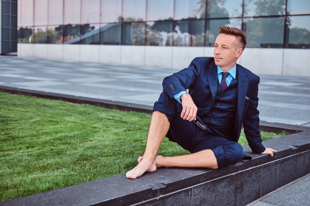 Ein Mann im Anzug sitzt barfüßig auf einer Mauer vor einem Geschäftsgebäude